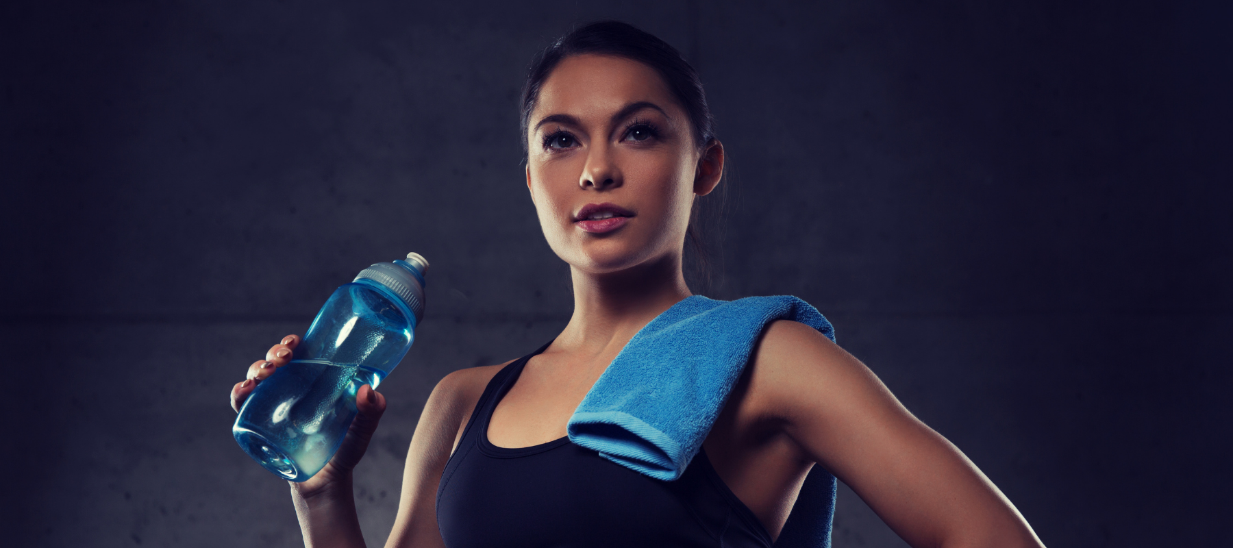 "Junge Frau in Sportkleidung hält blaue Wasserflasche und Handtuch, vor dunklem Hintergrund nach Workout."